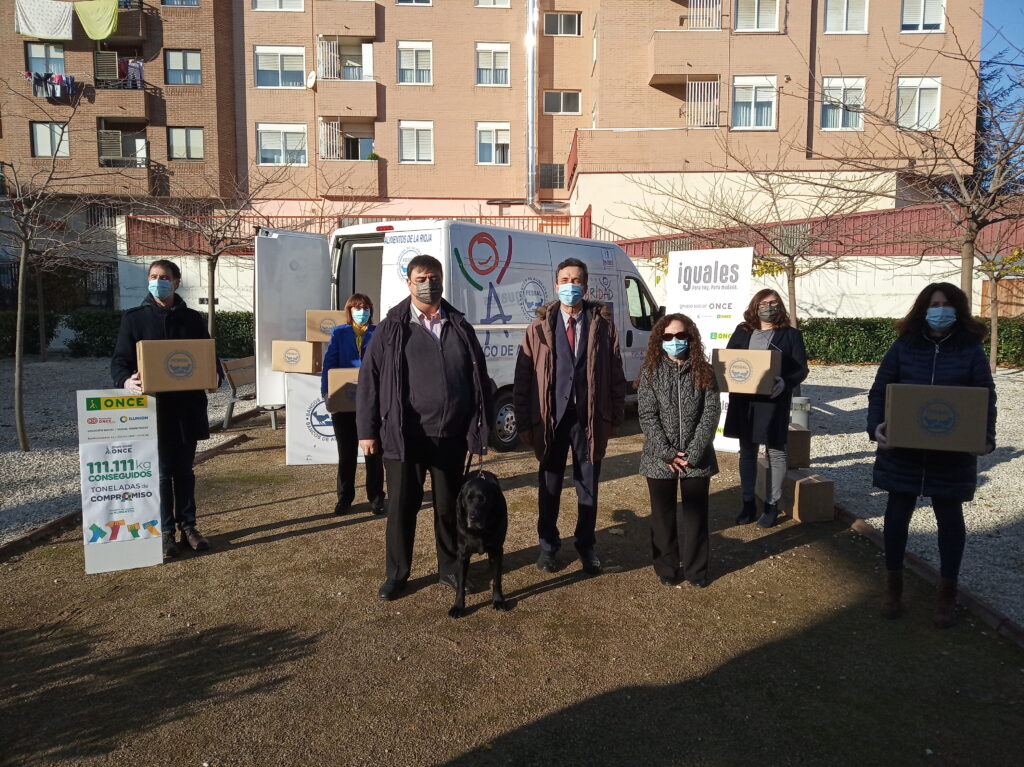 Grupo de personas con mascarilla junto a cajas de alimentos y una pancarta.